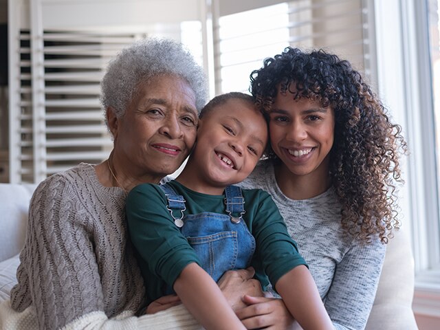 A beautiful senior woman sits on a couch with her granddaughter and daughter as they cuddle with each other and smile in this portrait..